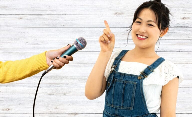 A cheerful woman in denim overalls raising her finger as if making a point, with a hand holding a microphone toward her.