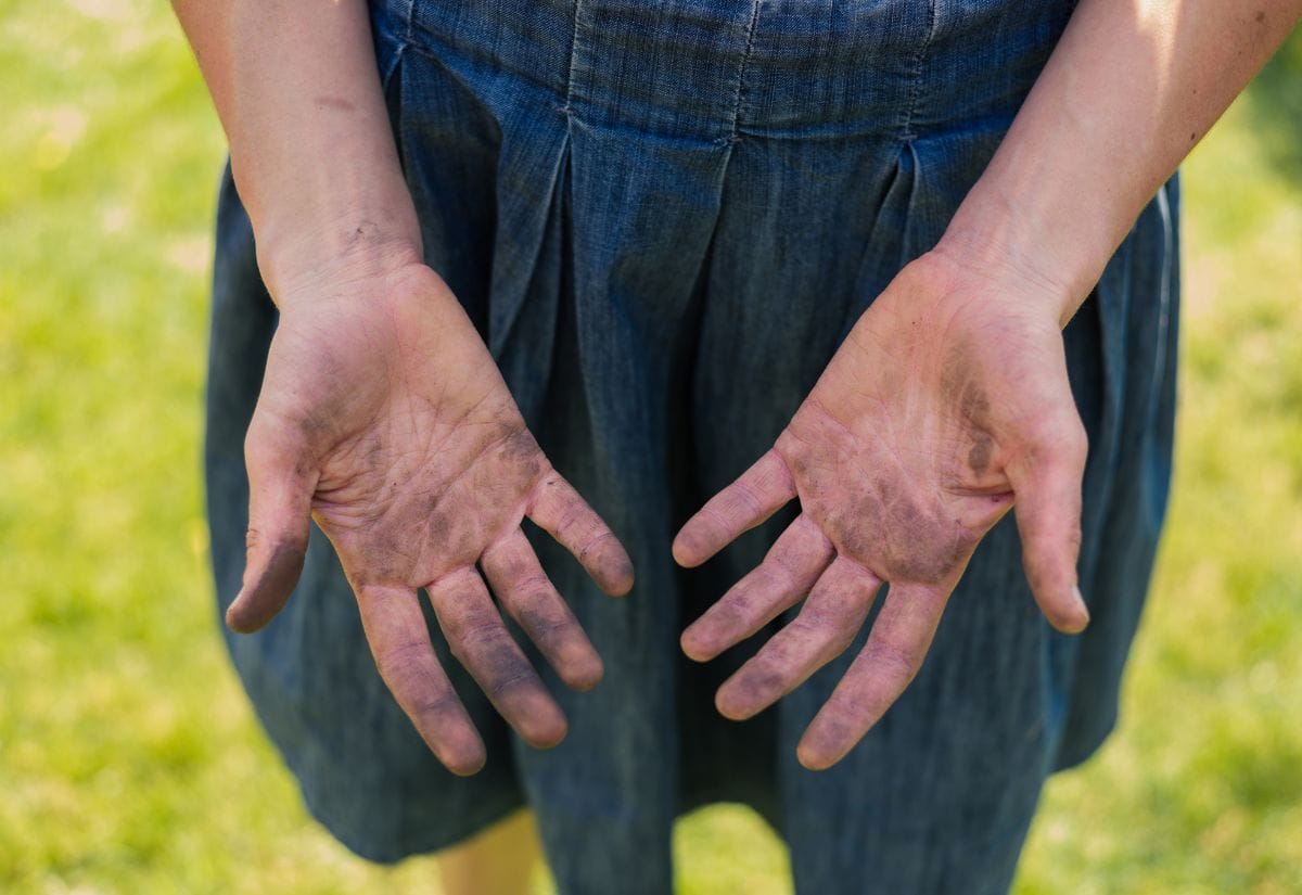 A close-up of a person’s hands covered in dirt, wearing a denim dress, standing on a grassy field.