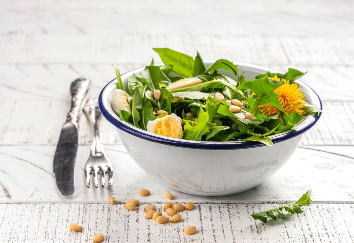 A rustic enamel bowl filled with foraged greens, dandelions, eggs, and pine nuts, placed on a white wooden table.