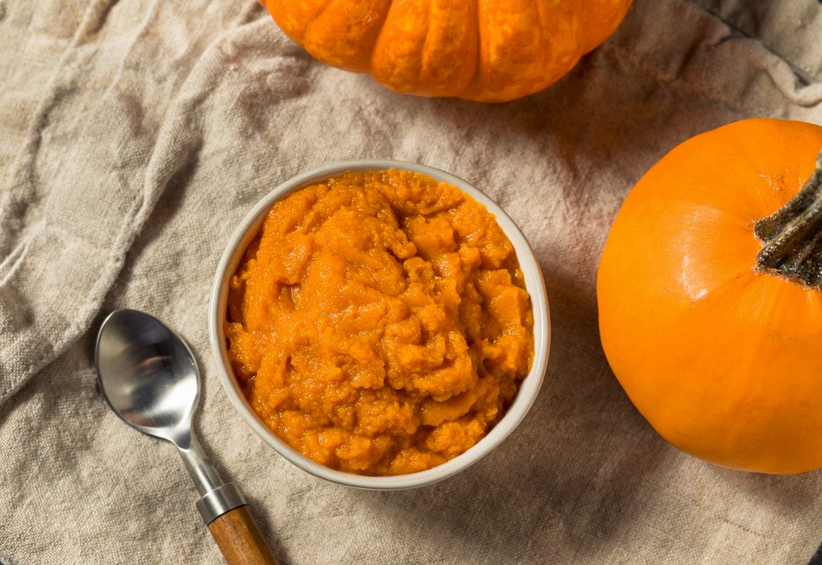 A small bowl of bright orange pumpkin puree sitting on a neutral linen cloth with two mini pumpkins and a spoon nearby, styled in soft natural light.