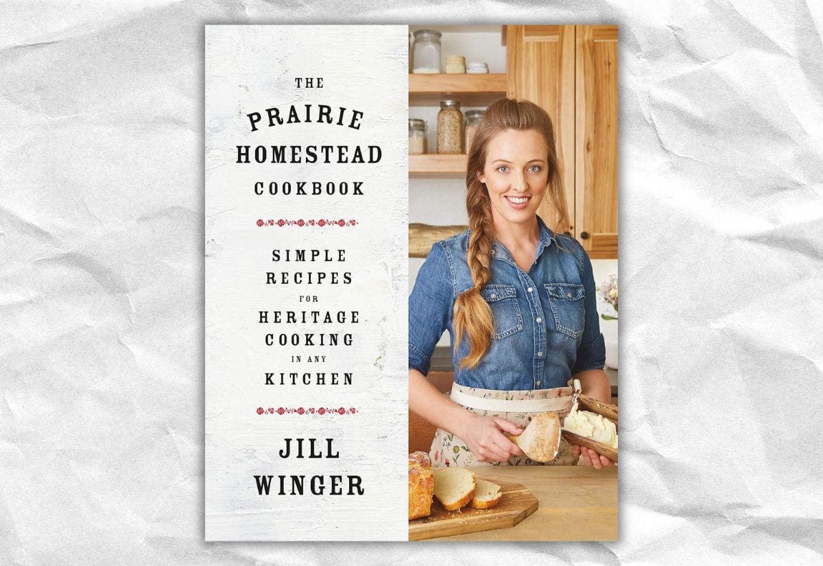 Cover of The Prairie Homestead Cookbook by Jill Winger, featuring Jill in a rustic kitchen slicing homemade bread.