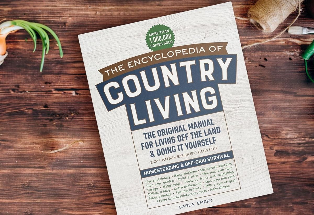 Cover of The Encyclopedia of Country Living by Carla Emery on a wooden table with rustic tools nearby.