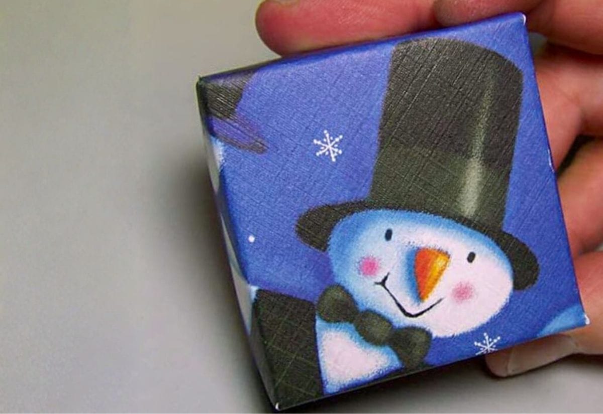 Close-up of a small gift box made from a recycled Christmas card featuring a smiling snowman in a black top hat, showing how to upcycle holiday cards into handmade packaging.