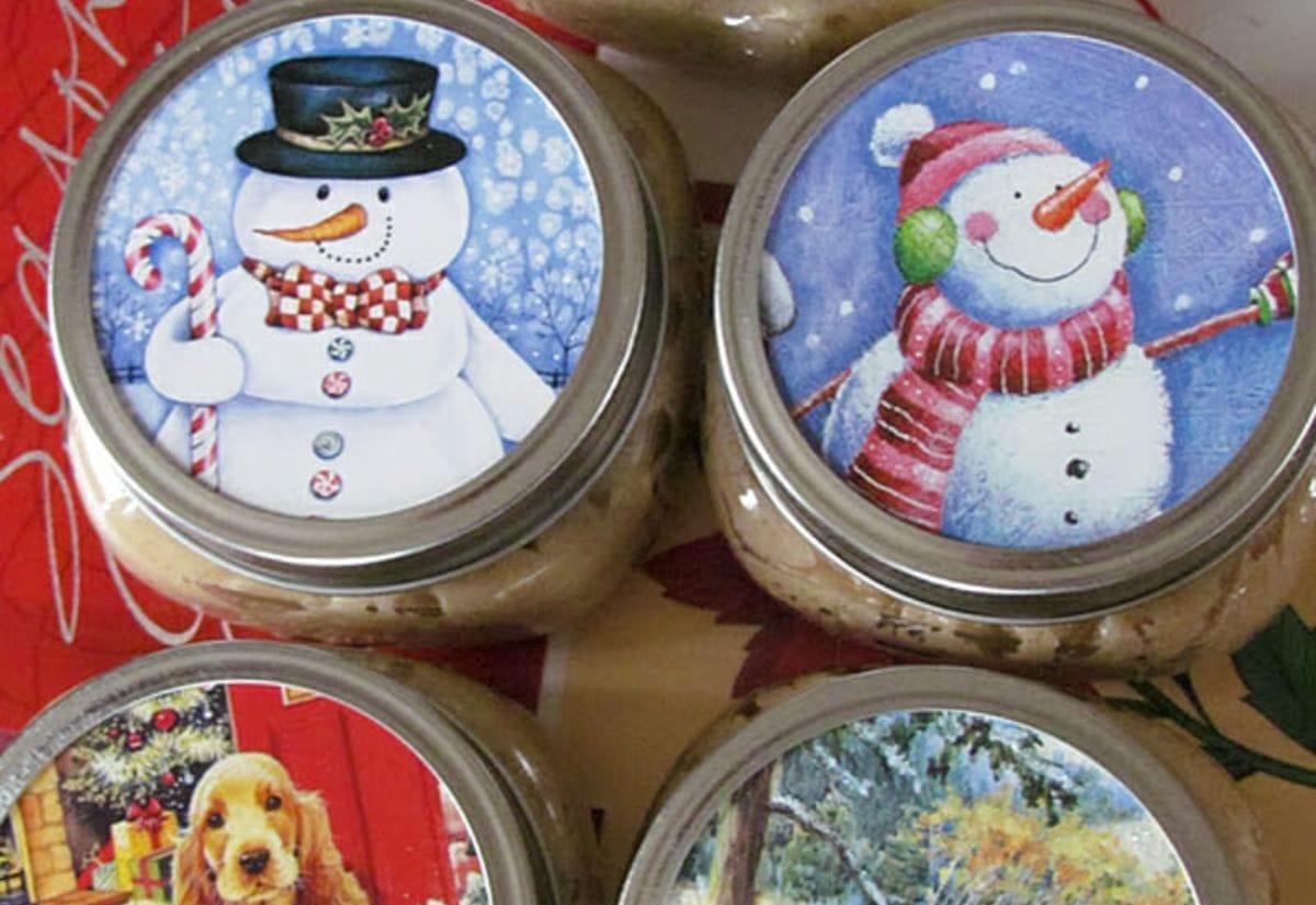 Four mason jars topped with recycled Christmas card cutouts of snowmen and winter scenes, showing how to turn old holiday cards into decorative jar lid covers for gifts.