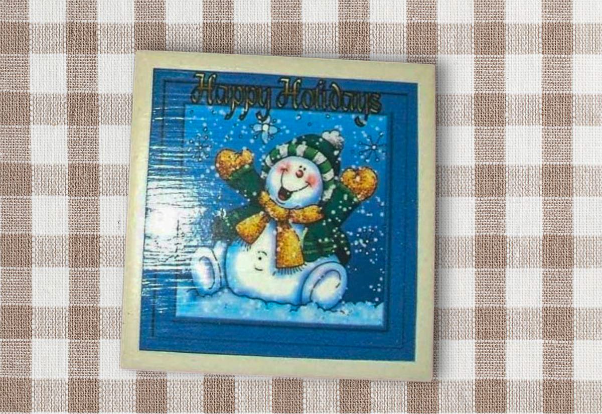 A square coaster made from a laminated “Happy Holidays” card with a cheerful snowman wearing a green hat and scarf, displayed on a tan gingham background.