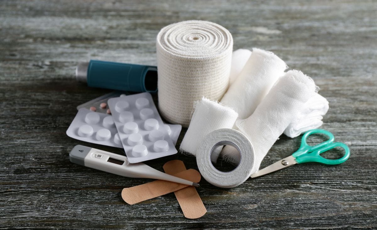 A collection of essential first aid supplies, including gauze, adhesive tape, scissors, thermometer, bandages, and medication, displayed on a rustic wooden surface."