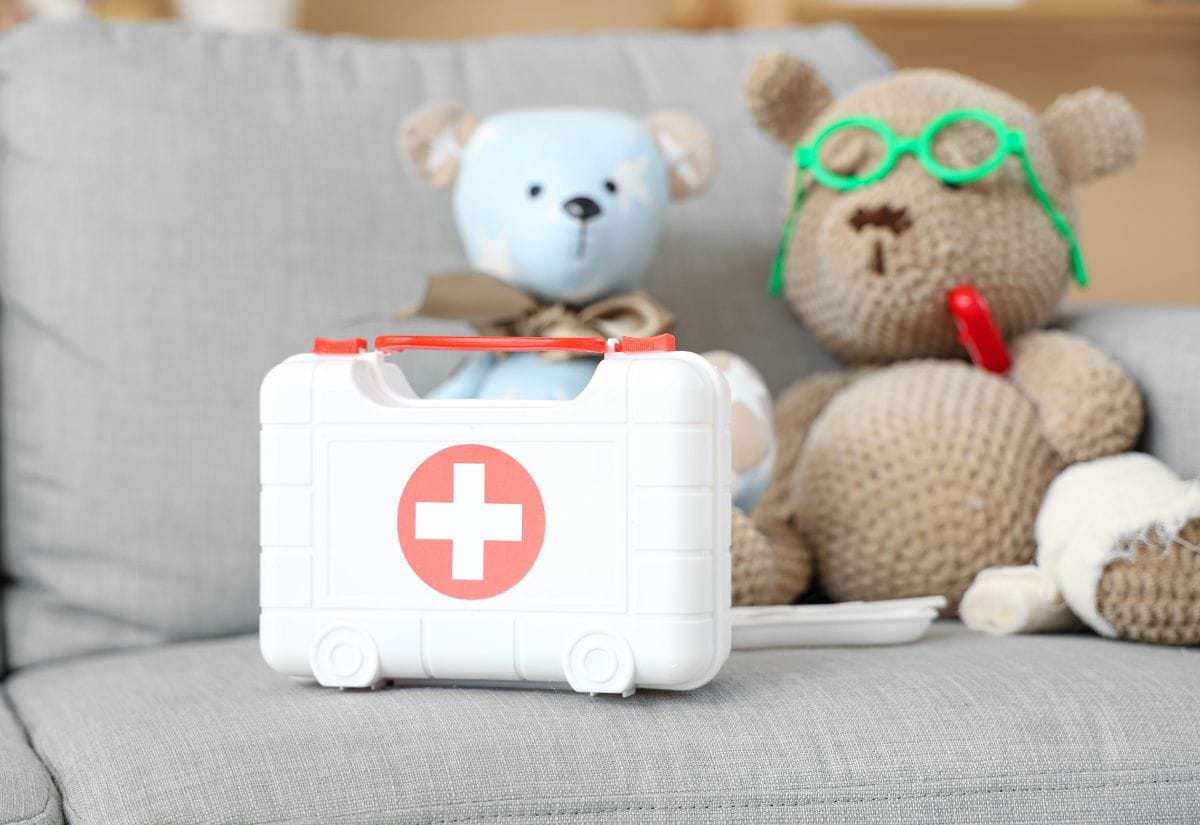 A compact first aid kit with a red cross symbol, placed on a couch alongside two teddy bears with playful accessories.