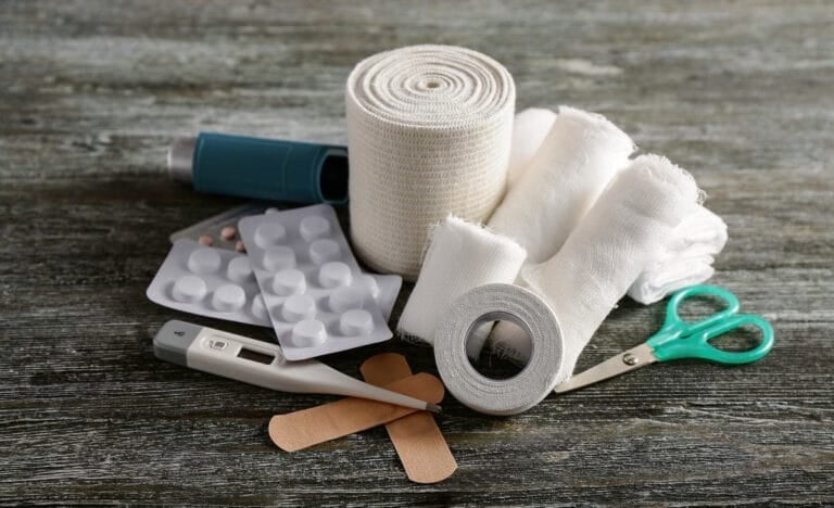 Assorted first aid items including gauze rolls, a digital thermometer, pills, adhesive bandages, and scissors arranged on a weathered wooden surface.