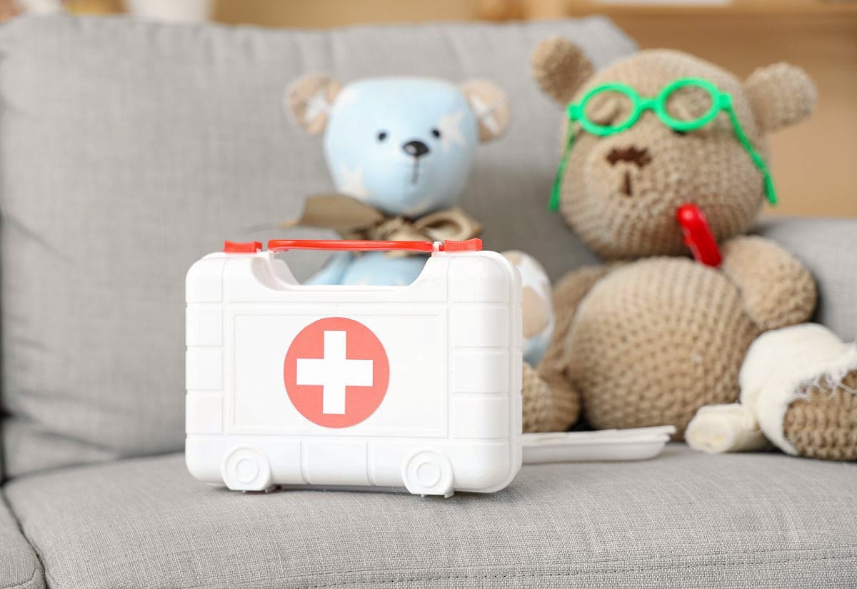 Plastic white first aid case with a red cross symbol placed on a couch next to two plush teddy bears, one wearing green toy glasses and a stethoscope.