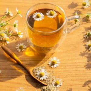 A clear glass mug filled with chamomile tea, adorned with chamomile flowers floating on the surface and scattered on a wooden table.