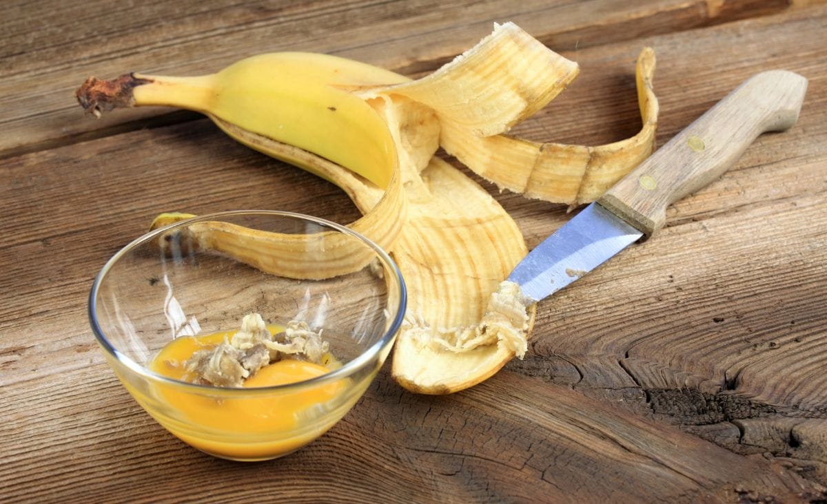 A ripe banana with its peel partially open, a rustic knife, and a glass bowl containing mashed banana and honey on a wooden surface.