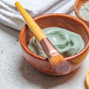 A wooden bowl of green clay facial mask with a wooden-handled brush resting on the edge, placed on a light countertop.