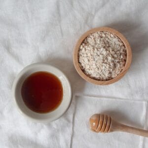 A small wooden bowl filled with rolled oats next to a small white bowl of honey with a honey dipper on a white cloth.