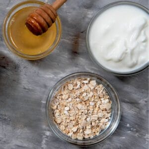 A small wooden bowl filled with rolled oats next to a small white bowl of honey with a honey dipper on a white cloth.