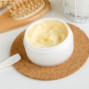 A white jar of thick, creamy moisturizer with a small spatula on a cork coaster beside a wooden hairbrush.