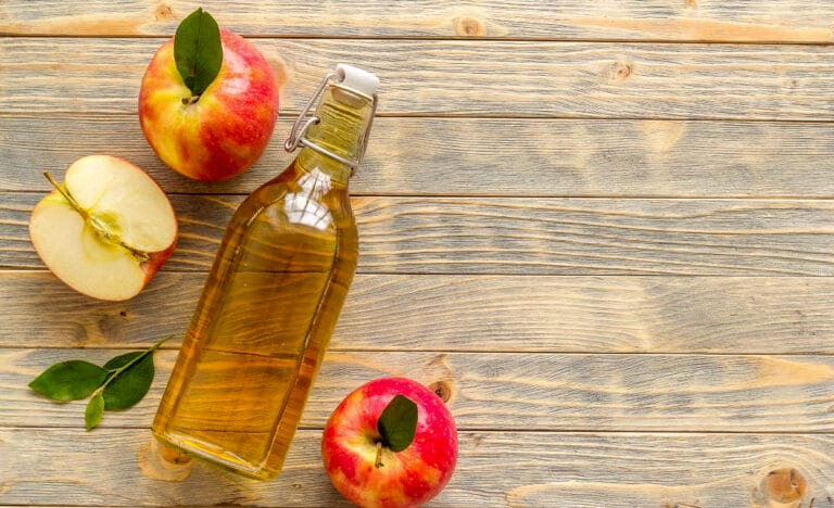A glass bottle of homemade hard apple cider on a wooden table, surrounded by red apples and apple slices.