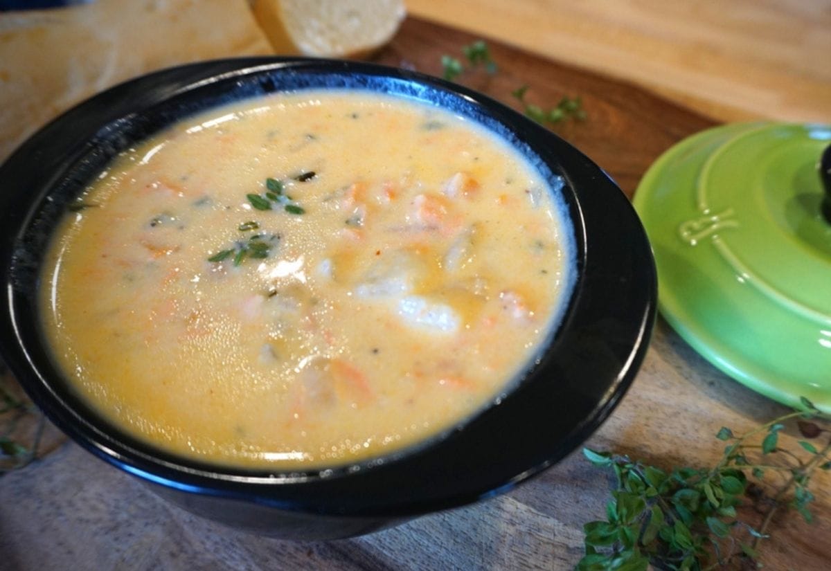 A black bowl filled with creamy salmon chowder garnished with fresh thyme, placed on a wooden surface with a green pot lid nearby.