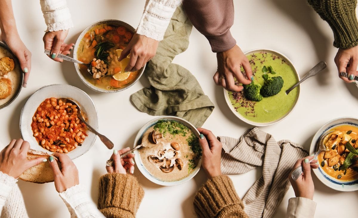 A cozy overhead view of multiple hands reaching for bowls of various soups, including bean stew, creamy mushroom soup, green broccoli soup, and squash soup, arranged on a table with bread and cloth napkins.