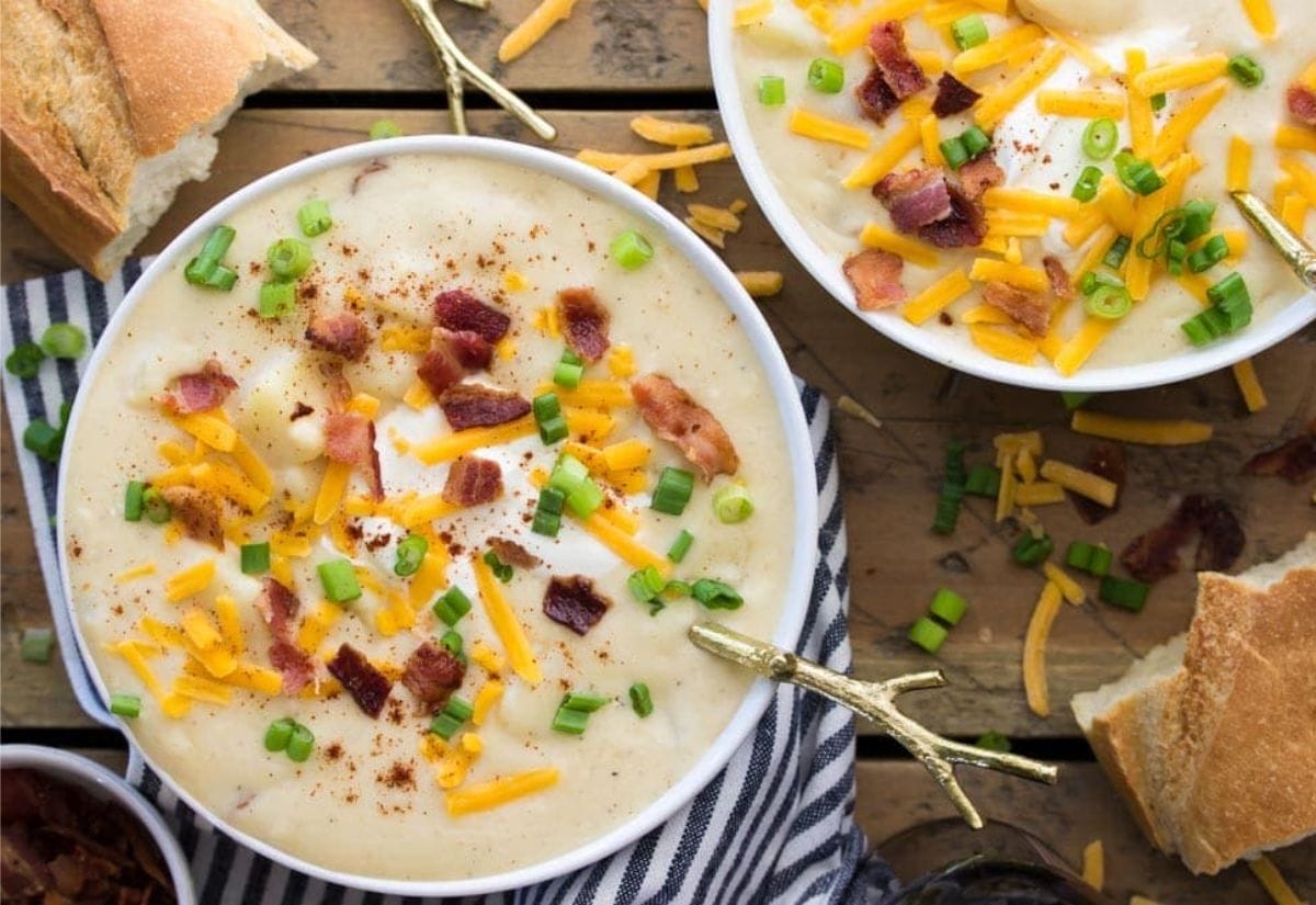 Two bowls of creamy loaded baked potato soup topped with shredded cheddar cheese, crispy bacon, and green onions, served with crusty bread.