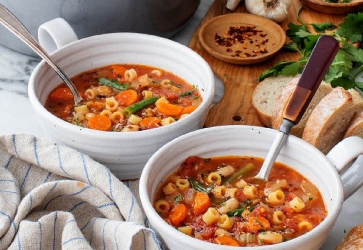 Two bowls of hearty minestrone soup filled with vegetables, pasta, and beans, served alongside crusty bread on a rustic wooden table.