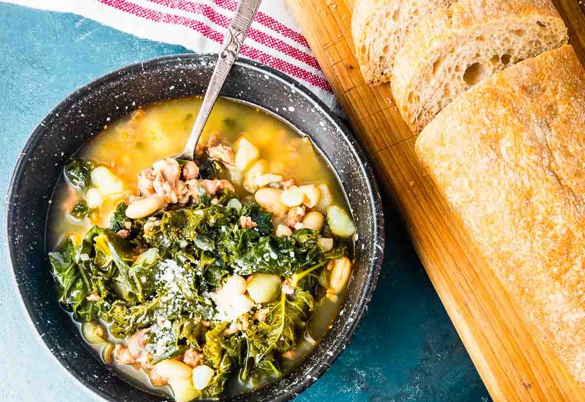 A hearty bowl of spicy sausage and kale soup with white beans, topped with grated Parmesan, served alongside slices of crusty bread on a wooden board.