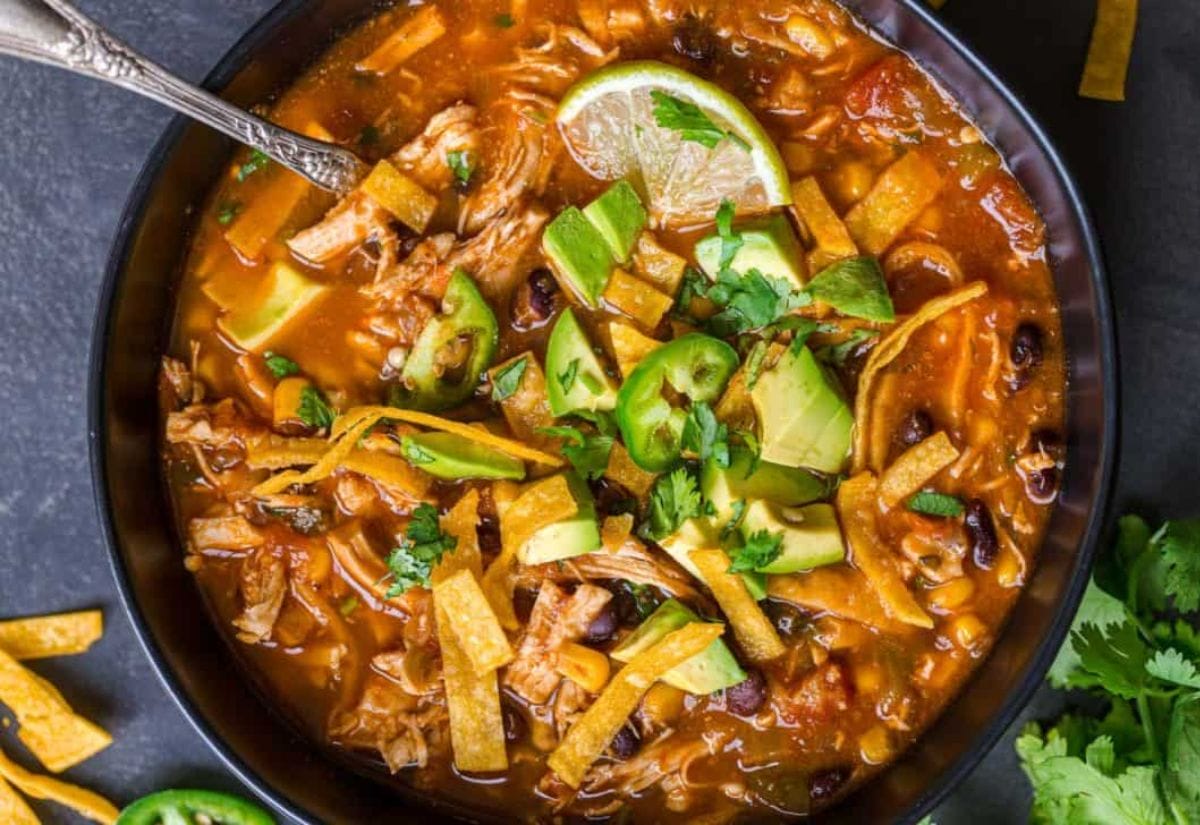 A hearty bowl of chicken tortilla soup topped with avocado chunks, crispy tortilla strips, jalapeño slices, fresh cilantro, and a wedge of lime.