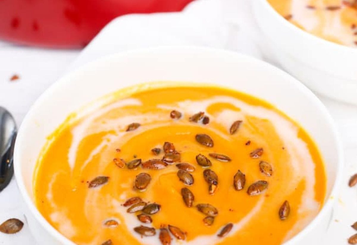 A creamy bowl of butternut squash and carrot soup topped with roasted pumpkin seeds and a swirl of cream, served in a white bowl.