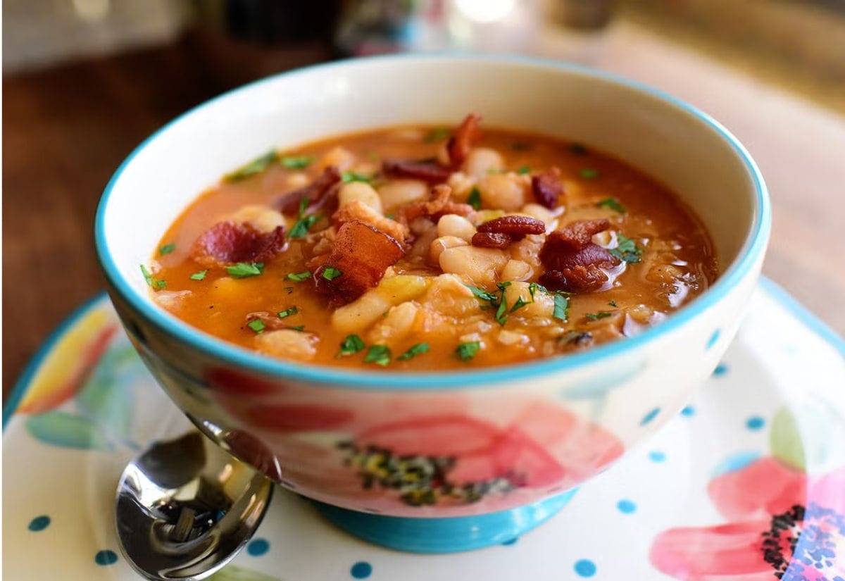 A colorful bowl of hearty bean and bacon soup, garnished with crispy bacon and fresh parsley, served on a floral-patterned plate with a spoon.