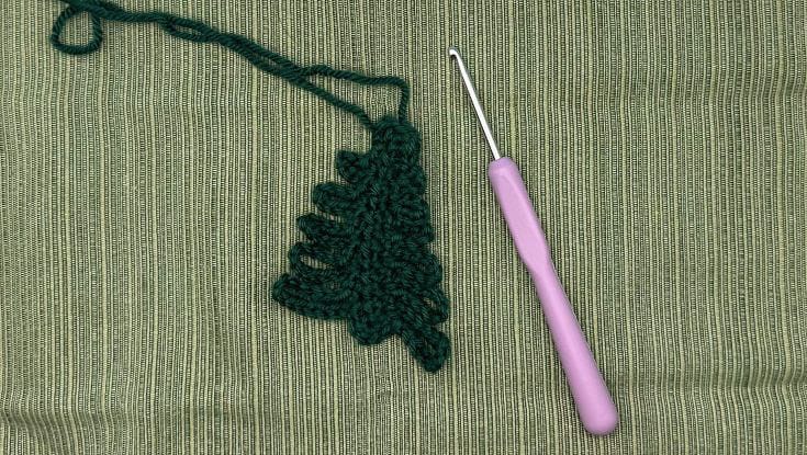 The nearly finished crochet Christmas tree ornament laid flat on a green background, showing all the loops formed down one side and the tree shape clearly visible beside a purple crochet hook.