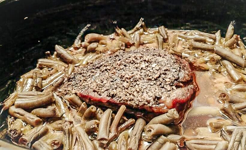 A pepper-seasoned beef roast cooking in a slow cooker with tender green beans and broth for a hearty winter meal.