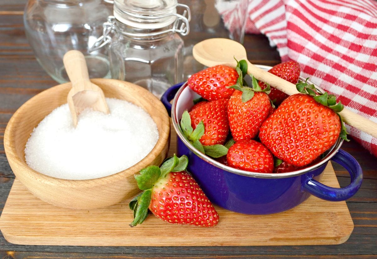 Fresh strawberries in a bowl, jars for canning, and a wooden scoop of sugar on a rustic wooden table.