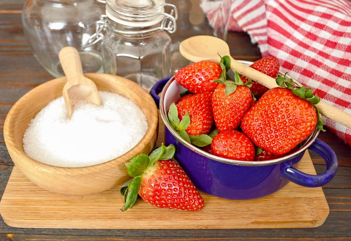 Fresh strawberries in a purple enamel pot beside a wooden bowl of granulated sugar. Perfect for a post comparing traditional and no-sugar jam methods.