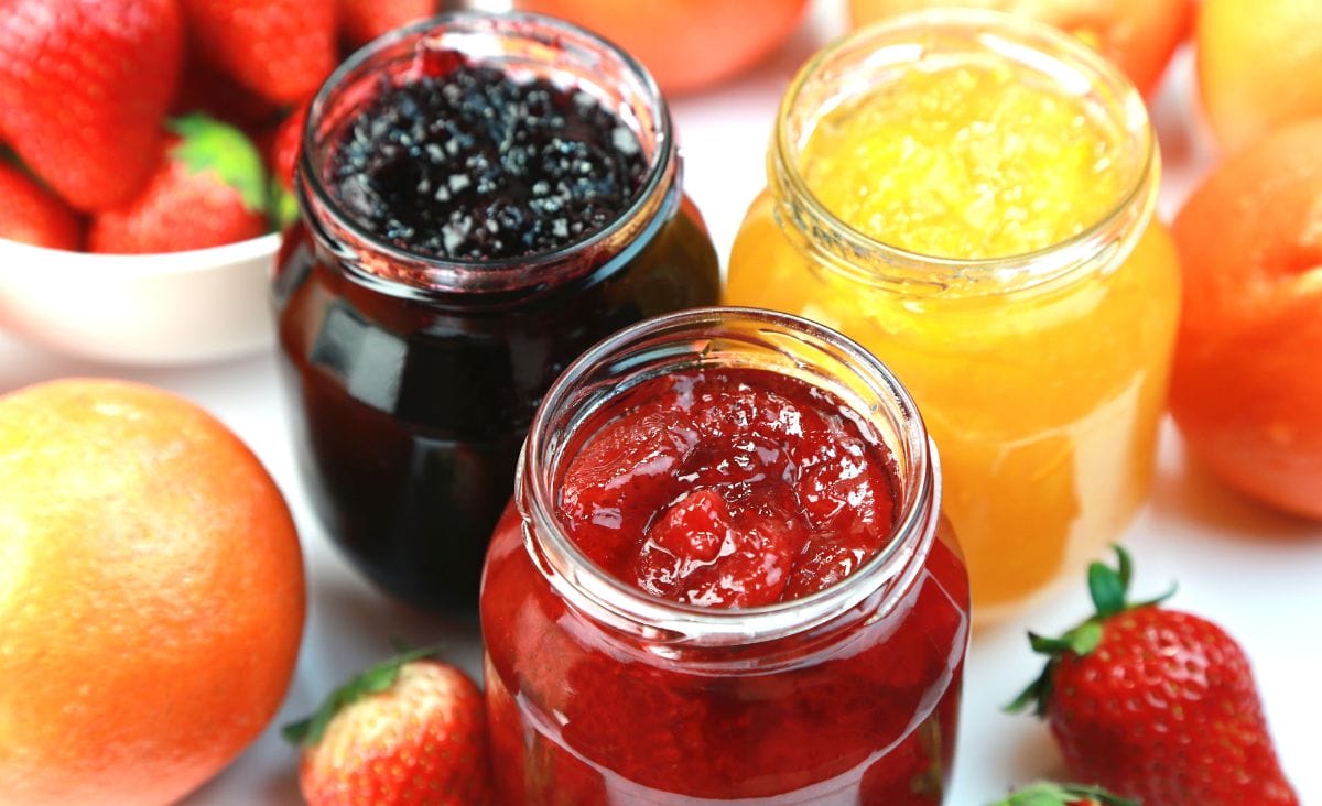 Three jars of jam in vibrant colors—blackberry, strawberry, and apricot—surrounded by fresh fruit on a bright table.