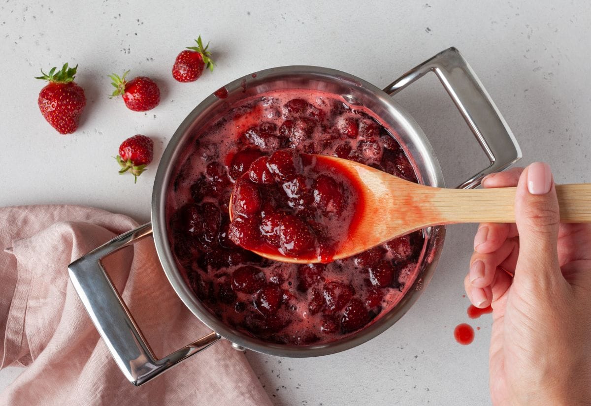 Cooking fresh strawberries in a pot to make jam, with a wooden spoon lifting the chunky mixture.