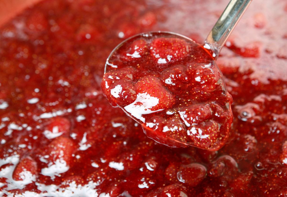 A ladle scooping thick strawberry jam with visible chunks of fruit, surrounded by a glossy red mixture.