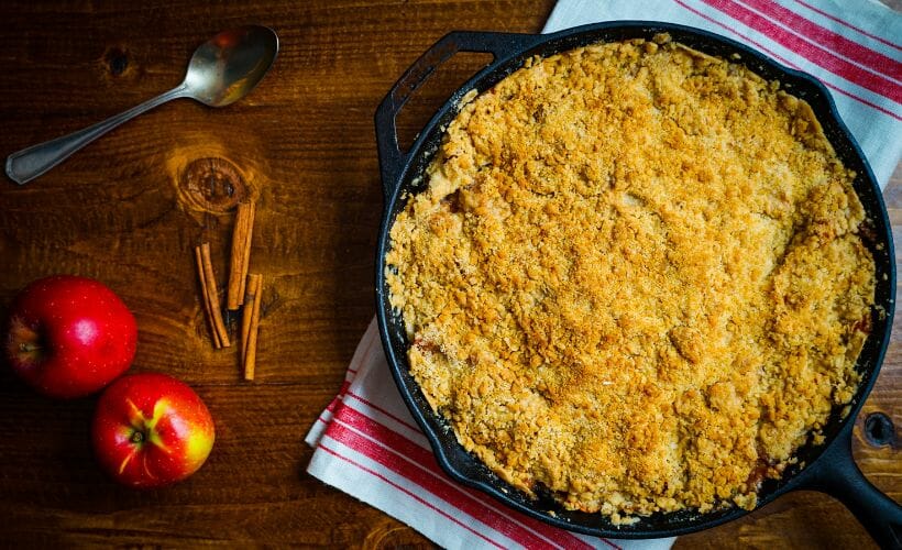 A cast iron pan of apple crumble laying on a wooden table with apples and cinnamon sticks.