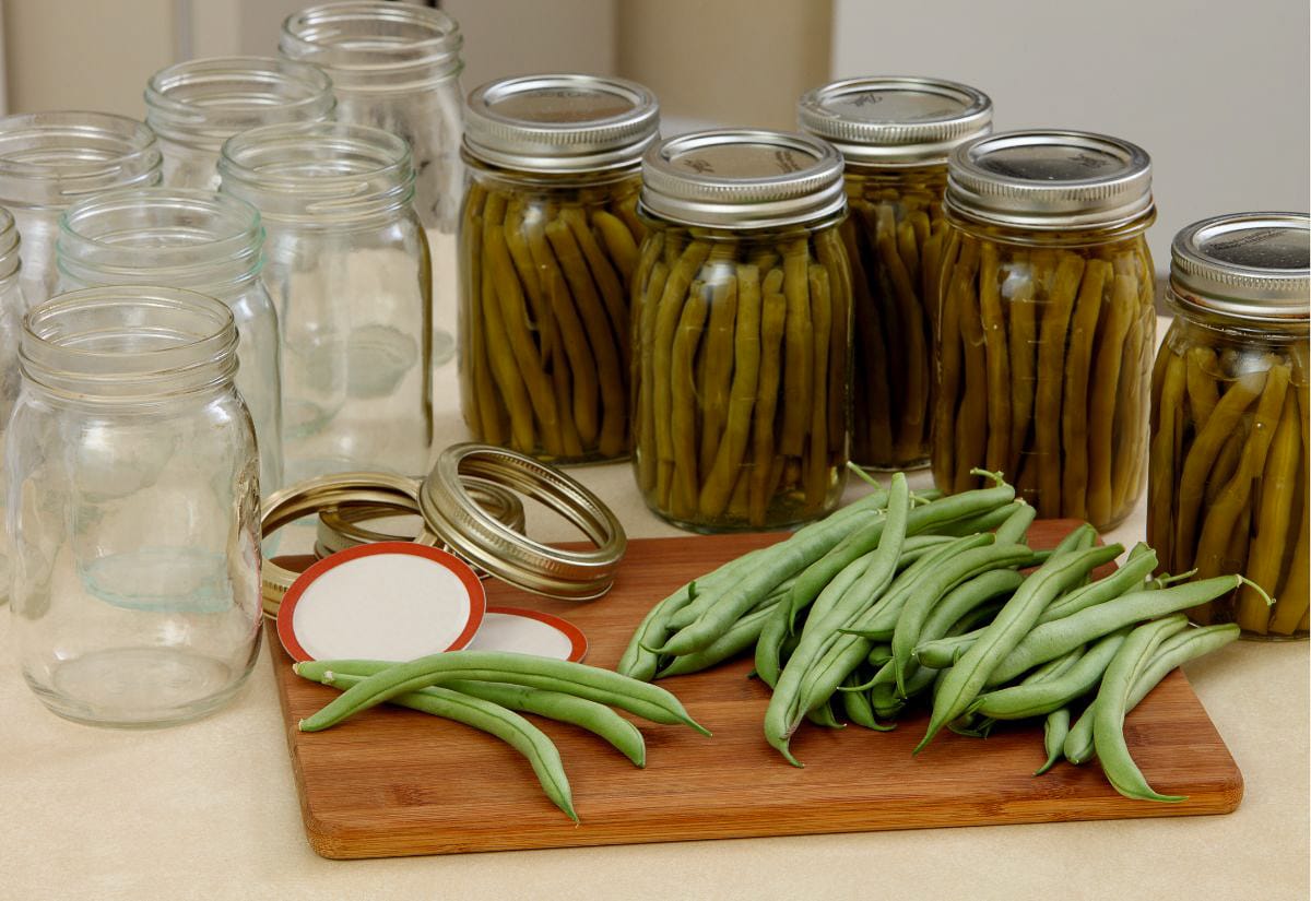 Fresh green beans on a cutting board next to sealed mason jars of home-canned beans and empty jars, showcasing food preservation on a modern homestead.