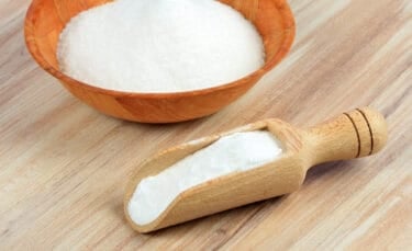Close-up of a wooden bowl and scoop filled with washing soda, sitting on a light wood surface.