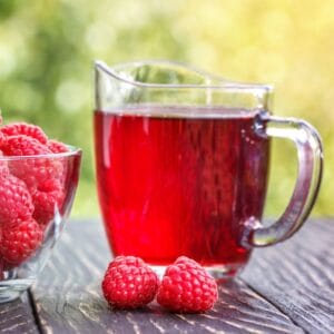 A pitcher of raspberry cordial and a bowl of fresh raspberries on a wooden table outdoors.