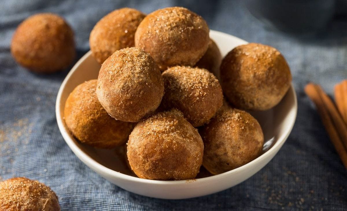 A white bowl filled with golden-brown sourdough donut holes dusted in cinnamon sugar, sitting on a blue cloth with a few scattered crumbs — a cozy, homemade fall treat.