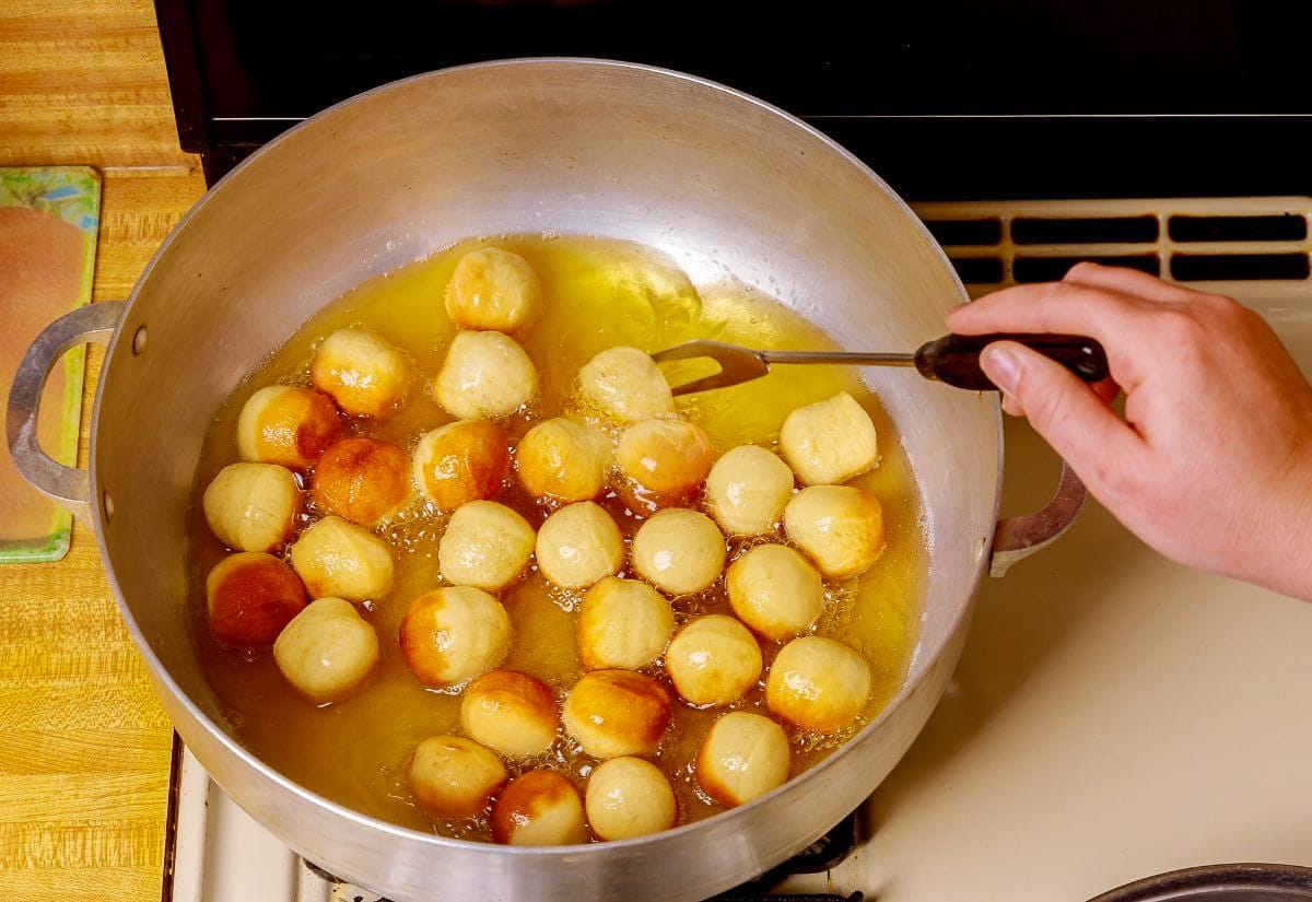 Freshly shaped donut holes frying in a large pot of oil on the stovetop, with one being turned by hand as they turn golden brown.