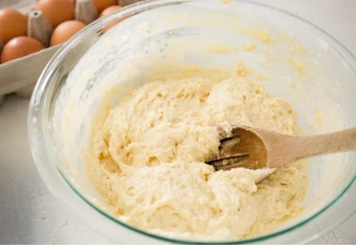 A glass bowl of sourdough donut dough being mixed with a wooden spoon, surrounded by eggs and baking ingredients on a kitchen counter.