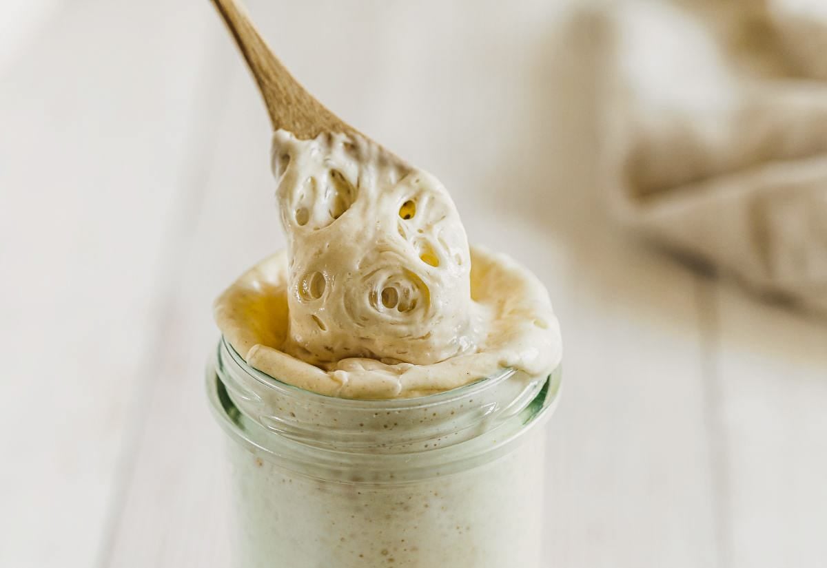 A bubbly, active sourdough starter stretching over the rim of a glass jar, showing its airy texture and strength — ready to bake with.