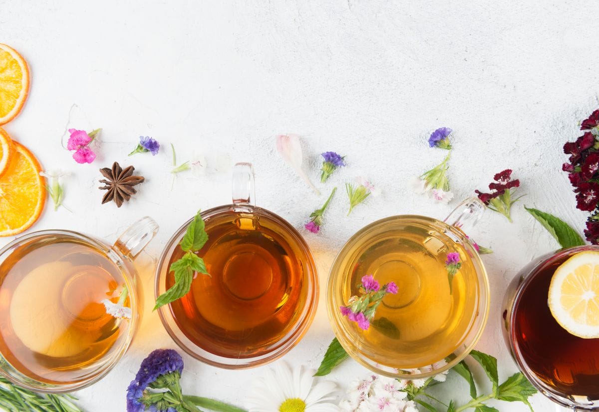 An assortment of colorful herbal teas in glass cups, garnished with fresh flowers and citrus slices on a bright background.