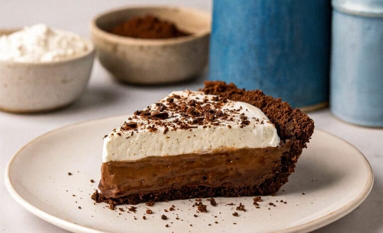 A slice of chocolate pudding pie with a chocolate crust and whipped cream topping on a white plate, with bowls of flour and cocoa powder and blue kitchen canisters blurred in the background.
