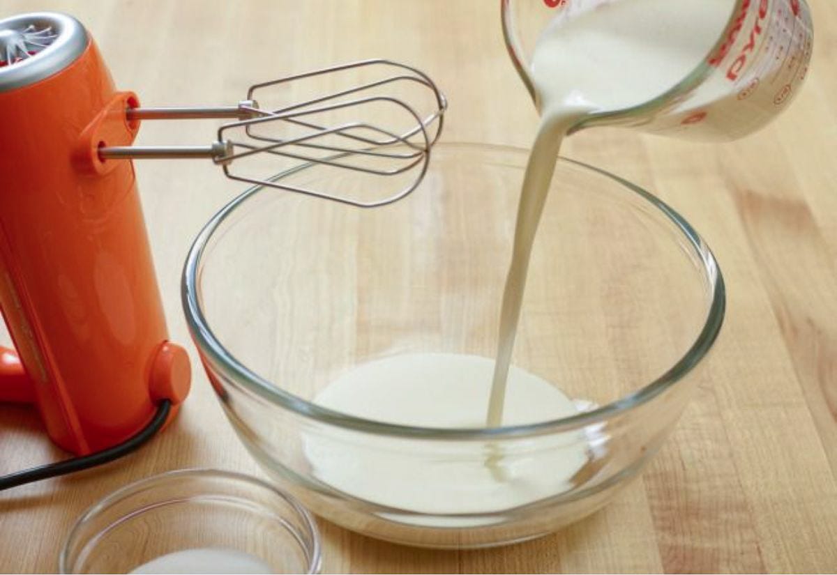 Heavy cream being poured into a glass mixing bowl beside an orange electric hand mixer, ready to be whipped for homemade whipped cream.