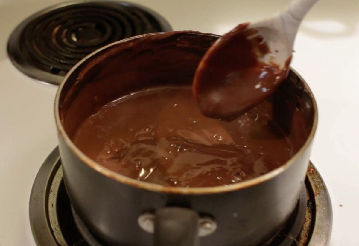 Homemade chocolate pudding thickening on the stovetop in a saucepan, with a spoon lifting the glossy pudding to show its texture.