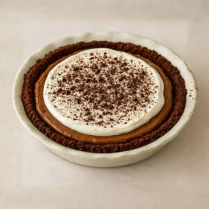 A full chocolate pudding pie with a homemade chocolate crust, silky chocolate filling, and a layer of whipped cream sprinkled with shaved chocolate, set in a white ceramic pie dish.