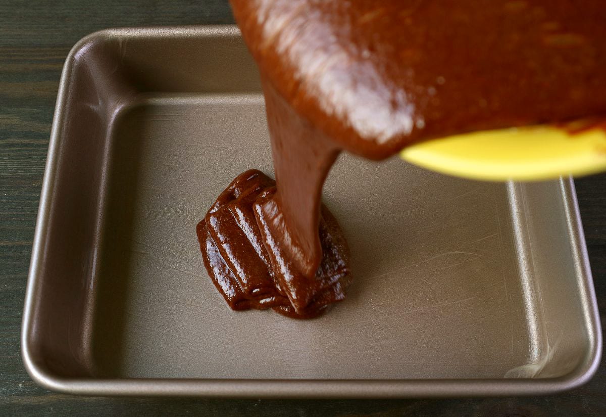 Melted chocolate fudge mixture being poured from a yellow bowl into a square metal baking pan.