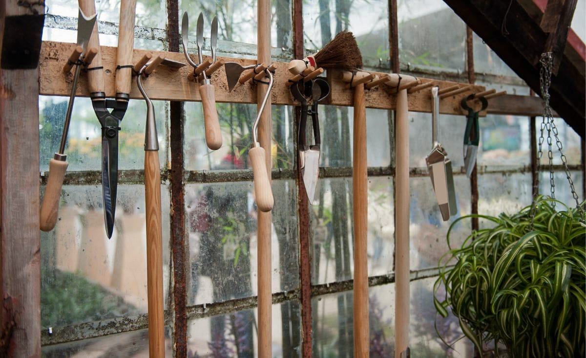 A collection of hand garden tools neatly hanging inside a greenhouse against an old glass window, showcasing essential gardening supplies in a rustic setting.